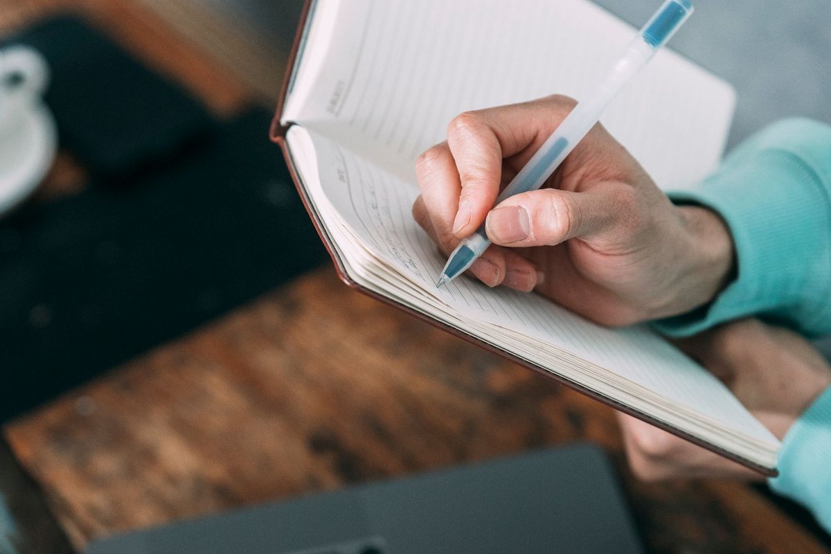 A hand writing notes in a notebook beside a laptop